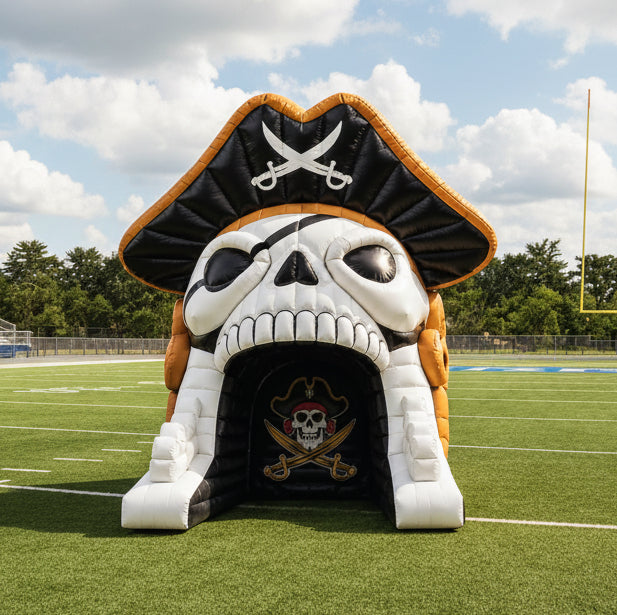 A pirate skull inflatable slide or tunnel set on a football field at night under stadium lights. The skull has a black bandana with crossed swords on top, white skull face, and gold trim, with a dark tunnel opening below.