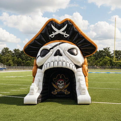 A pirate skull inflatable slide or tunnel set on a football field at night under stadium lights. The skull has a black bandana with crossed swords on top, white skull face, and gold trim, with a dark tunnel opening below.
