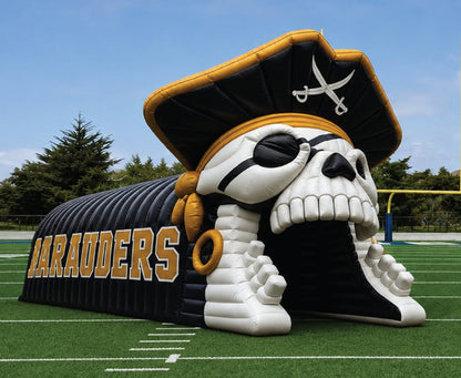An inflatable pirate skull tunnel designed for football, featuring a large skull with a black eye patch and a black pirate hat with gold trim, situated on a green turf field with white yard lines and a blue sky background.