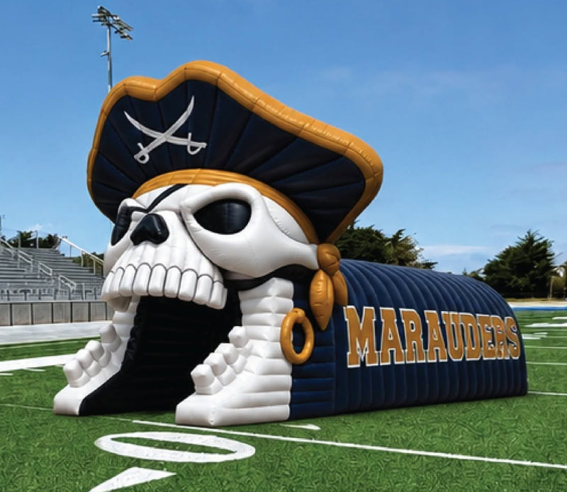A large pirate-themed inflatable tunnel on a green football field under a blue sky. The tunnel features a white skull wearing a black and gold pirate hat, with the word 'MARAUDERS' in gold on a dark blue body.